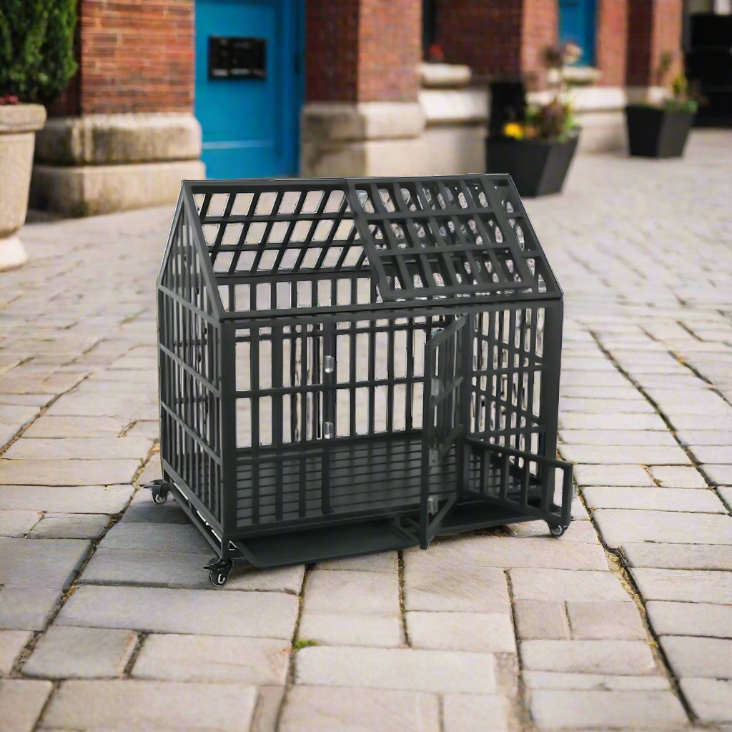 A black metal dog crate shaped like a house, featuring a door and wheels, placed on a cobblestone street with greenery in the background.